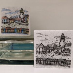 Two handmade ceramic tiles featuring illustrated seaside scenes with a lighthouse, buildings and beach huts.