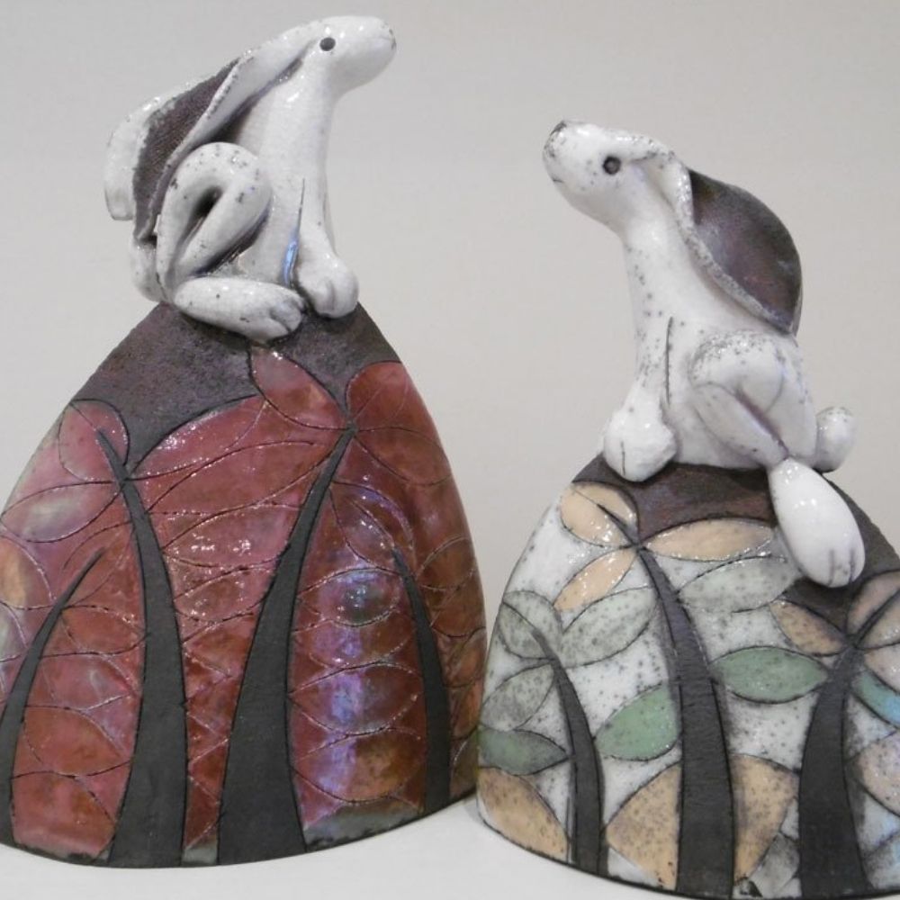 Two handmade ceramic hare sculptures sitting on patterned leaf-shaped bases.