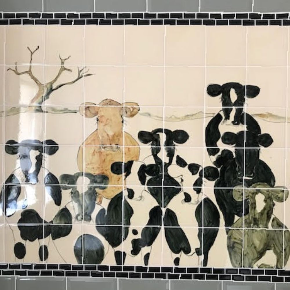 Decorative tiled wall mural featuring illustrated cows in a countryside scene with a black mosaic border.