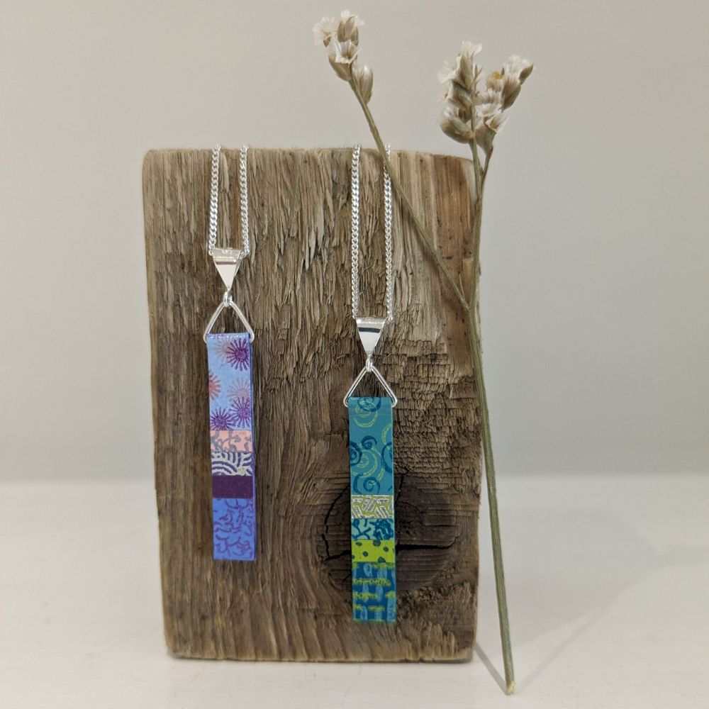 Two handmade rectangular pendant necklaces with colourful patterned designs displayed on rustic wood with dried flowers.