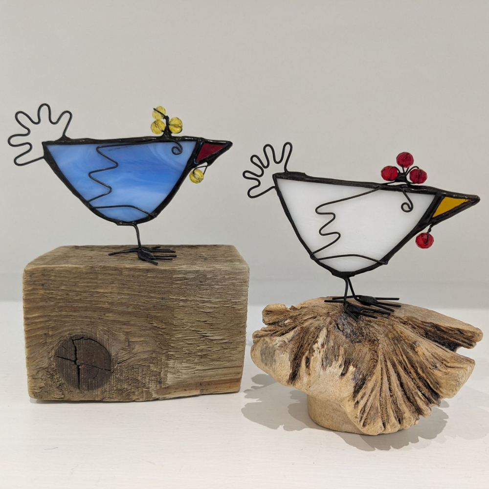 Handmade stained glass bird decorations in blue and white with wire details, berries and yellow accents, displayed on natural wood.