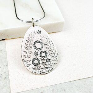 Silver pendant necklace with engraved floral and leafy botanical designs displayed on a white surface.