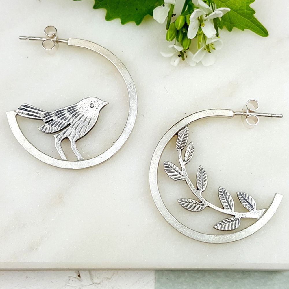 Pair of silver hoop-style earrings with an asymmetrical bird and leafy branch design, displayed on a white surface.