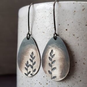 Pair of silver oval drop earrings with cut-out leaf designs, hanging from dark hook wires against a speckled ceramic background.