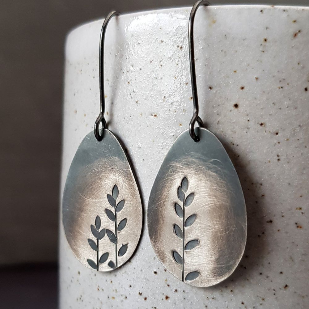 Pair of silver oval drop earrings with cut-out leaf designs, hanging from dark hook wires against a speckled ceramic background.