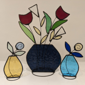 Handmade stained glass decorations featuring a dark blue flower vase with red and white flowers, beside yellow and blue fruit designs.
