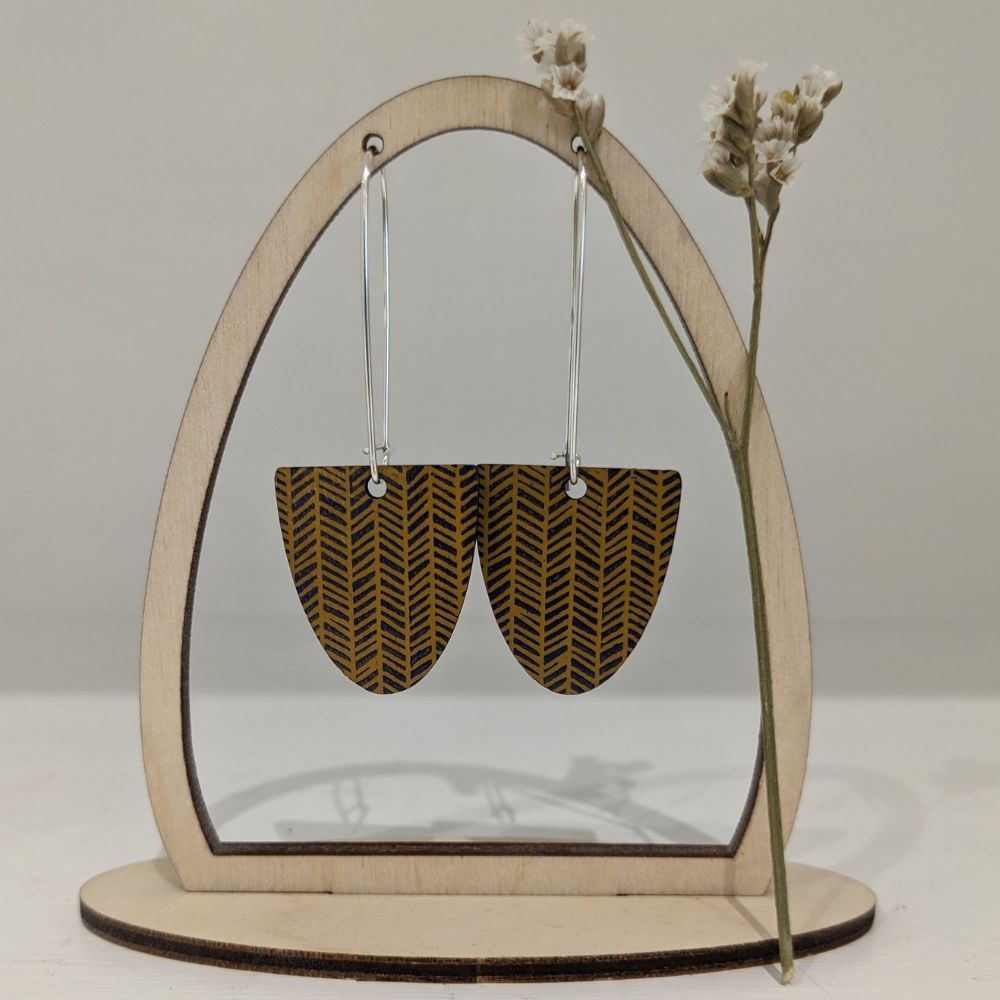 Pair of mustard yellow half-circle drop earrings with black chevron pattern, displayed on a wooden jewellery stand with dried flowers.