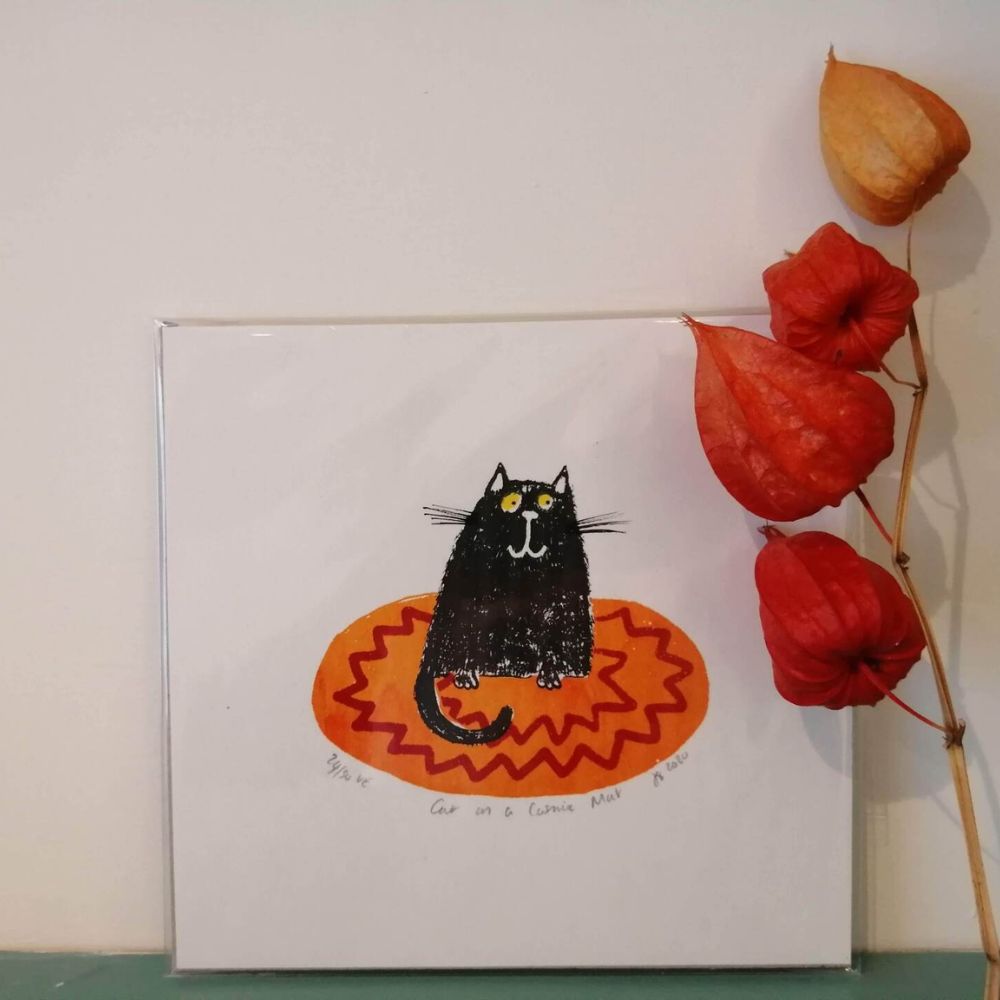 Art card featuring a black cat sitting on an orange patterned mat, displayed beside dried orange flowers.