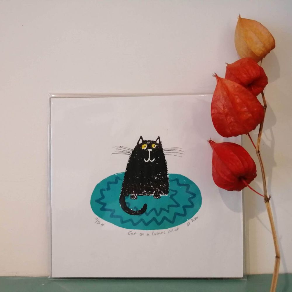 Art card featuring a black cat sitting on a blue patterned mat, displayed beside dried orange flowers.