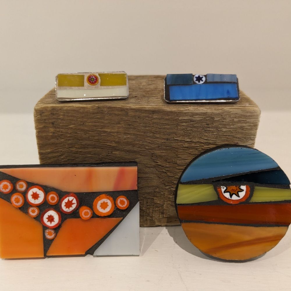 Display of handmade fused glass jewellery with colourful geometric designs, including stud earrings, brooches and statement pieces on rustic wood.