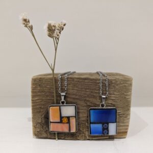 Two handmade square pendant necklaces with colourful geometric designs displayed on rustic wood with dried flowers.