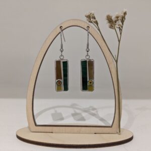 Pair of handmade rectangular drop earrings with green, brown and yellow details displayed on a wooden jewellery stand with dried flowers.